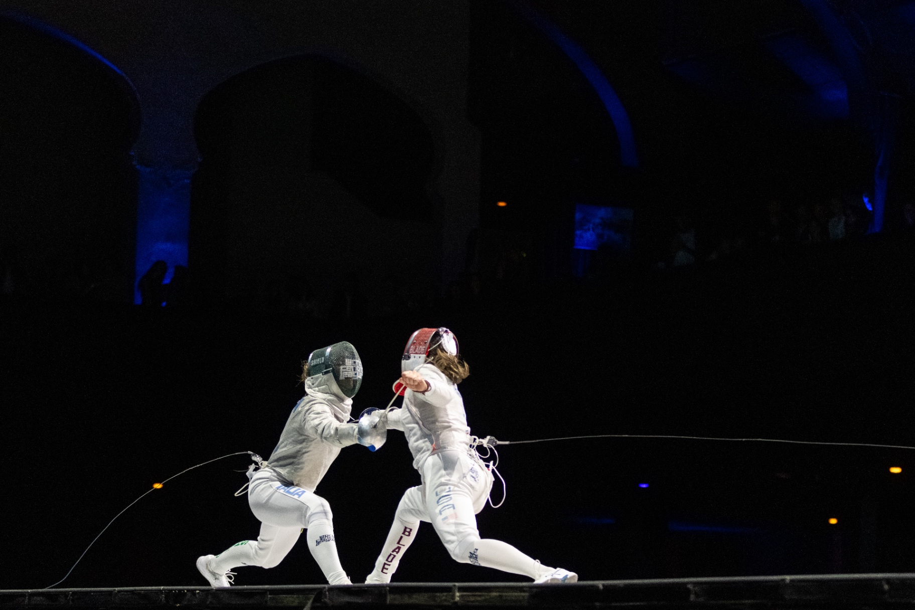 A Blade Collective fencer lunging on the piste, lit against deep blue arches.