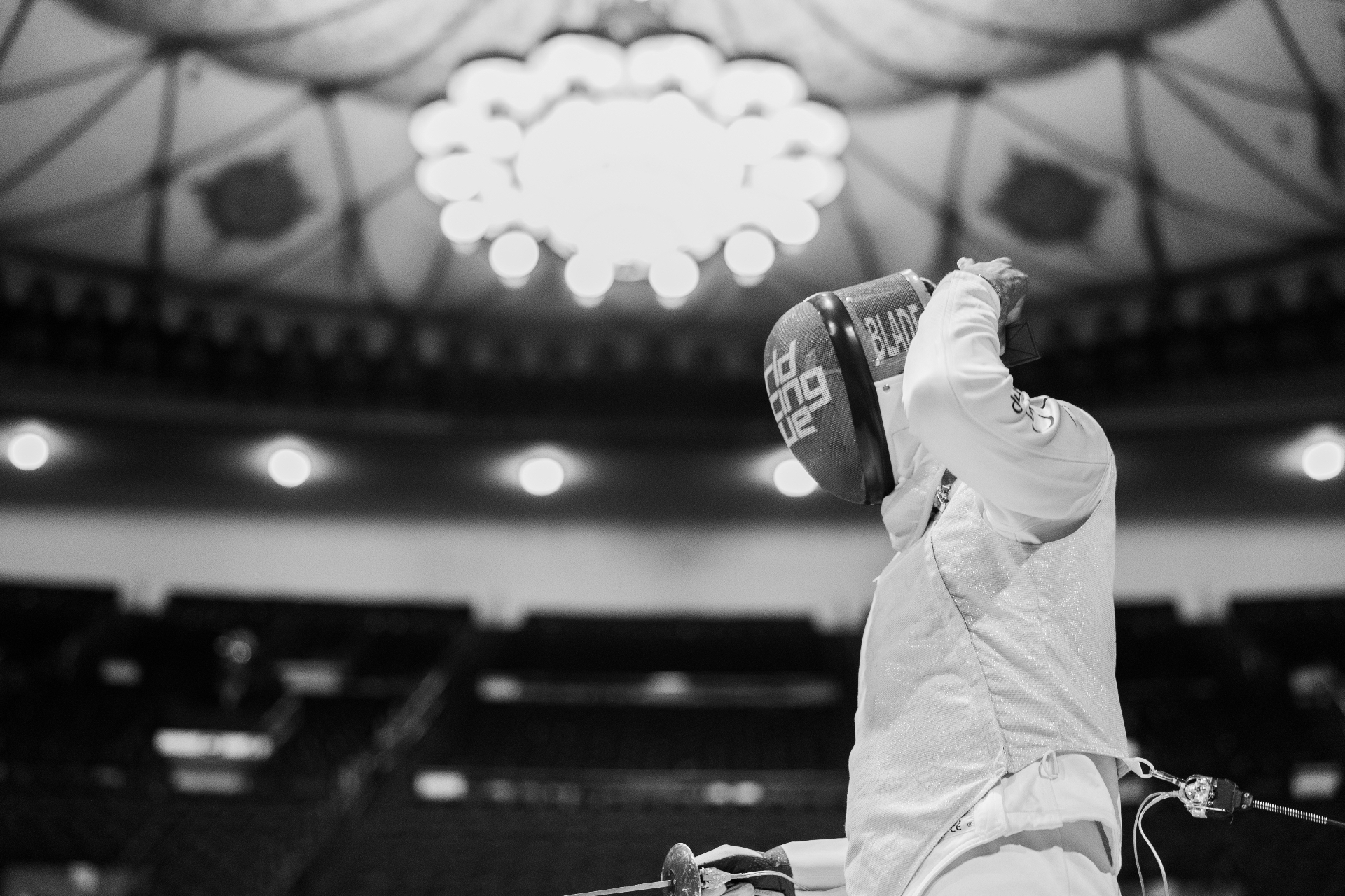 A fencer in a BLADE-marked mask raises a sabre under an ornate arena chandelier.