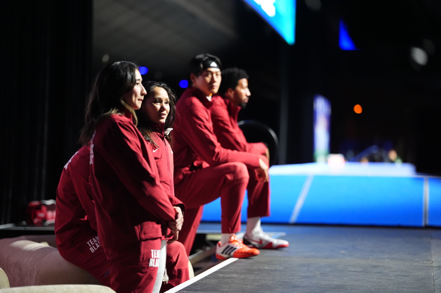 Team Blade athletes in deep red tracksuits seated at the side of the piste between bouts.