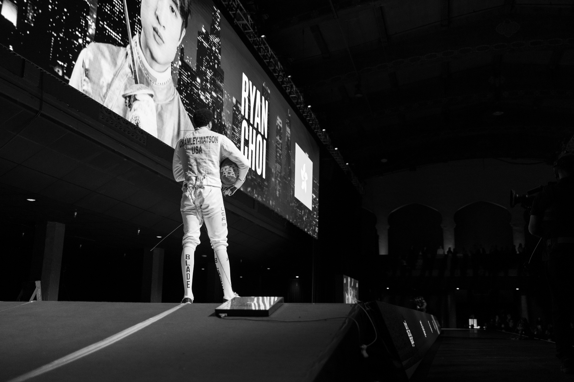 A Team Blade fencer walks toward the piste with a teammate's portrait projected on the arena screen behind.
